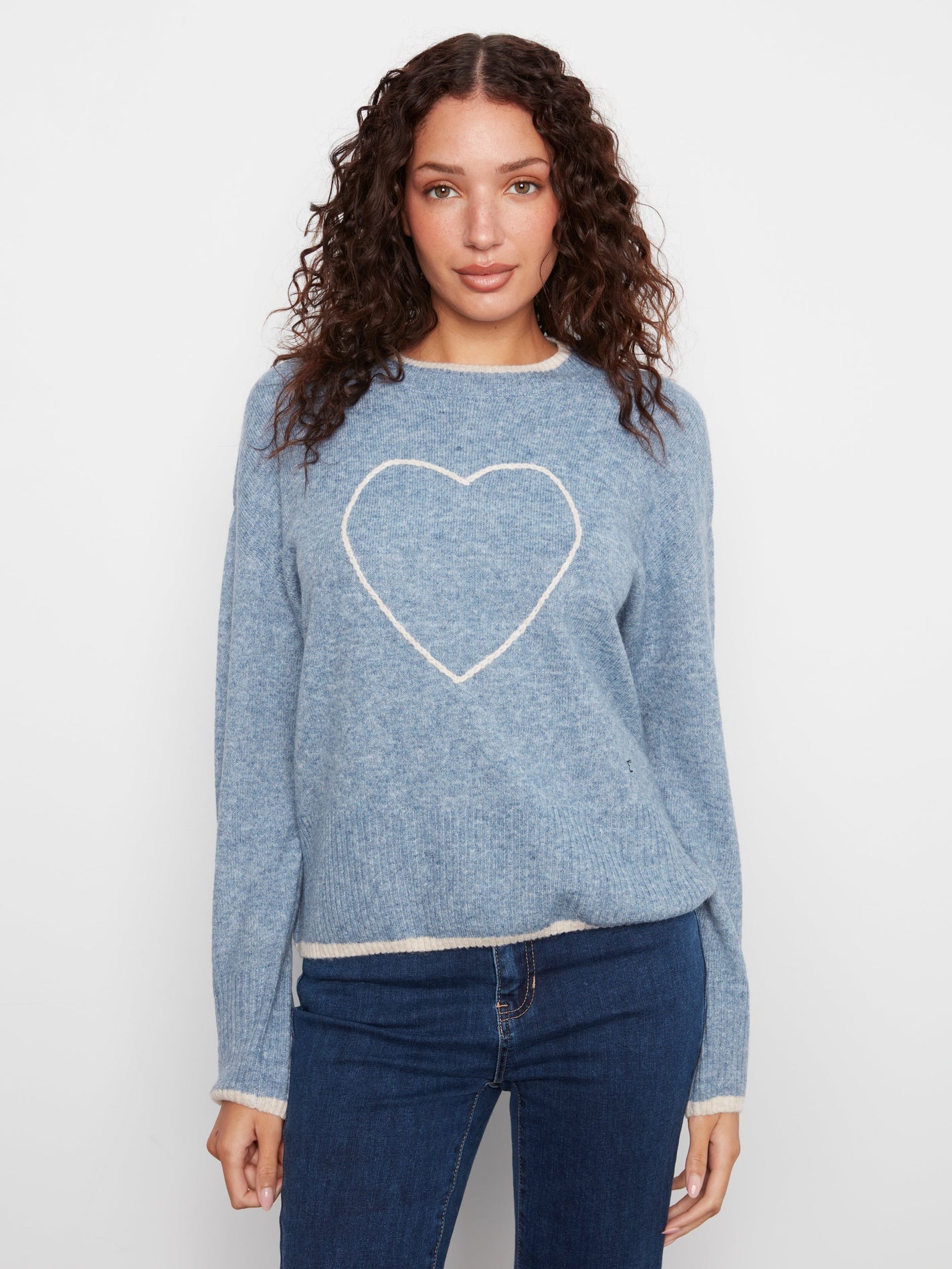 A woman with curly brown hair wearing a light blue knit sweater with a white heart outline on the chest, paired with dark blue jeans, against a plain light gray background.