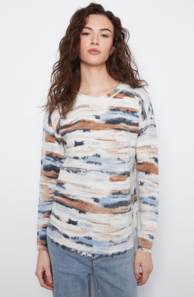 A woman modeling a long-sleeve knit sweater with an abstract watercolor pattern in beige, brown, blue, and white, paired with light blue jeans against a plain white background.