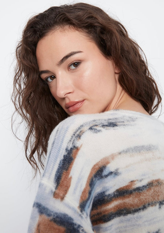 A close-up shot of a woman with wavy brown hair looking over her shoulder, wearing a fuzzy knit sweater with a blue, brown, and white abstract pattern against a plain white background.