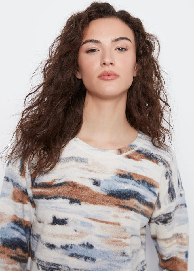 A woman with long, wavy brown hair wearing a fuzzy long-sleeve top with an abstract watercolor pattern in beige, brown, blue, and white, set against a plain white background.