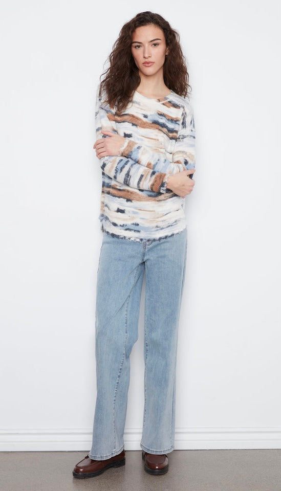 A woman modeling a long-sleeve knit sweater with a distressed abstract pattern in beige, brown, and blue, paired with light-wash wide-leg jeans and dark brown shoes, standing against a white wall.