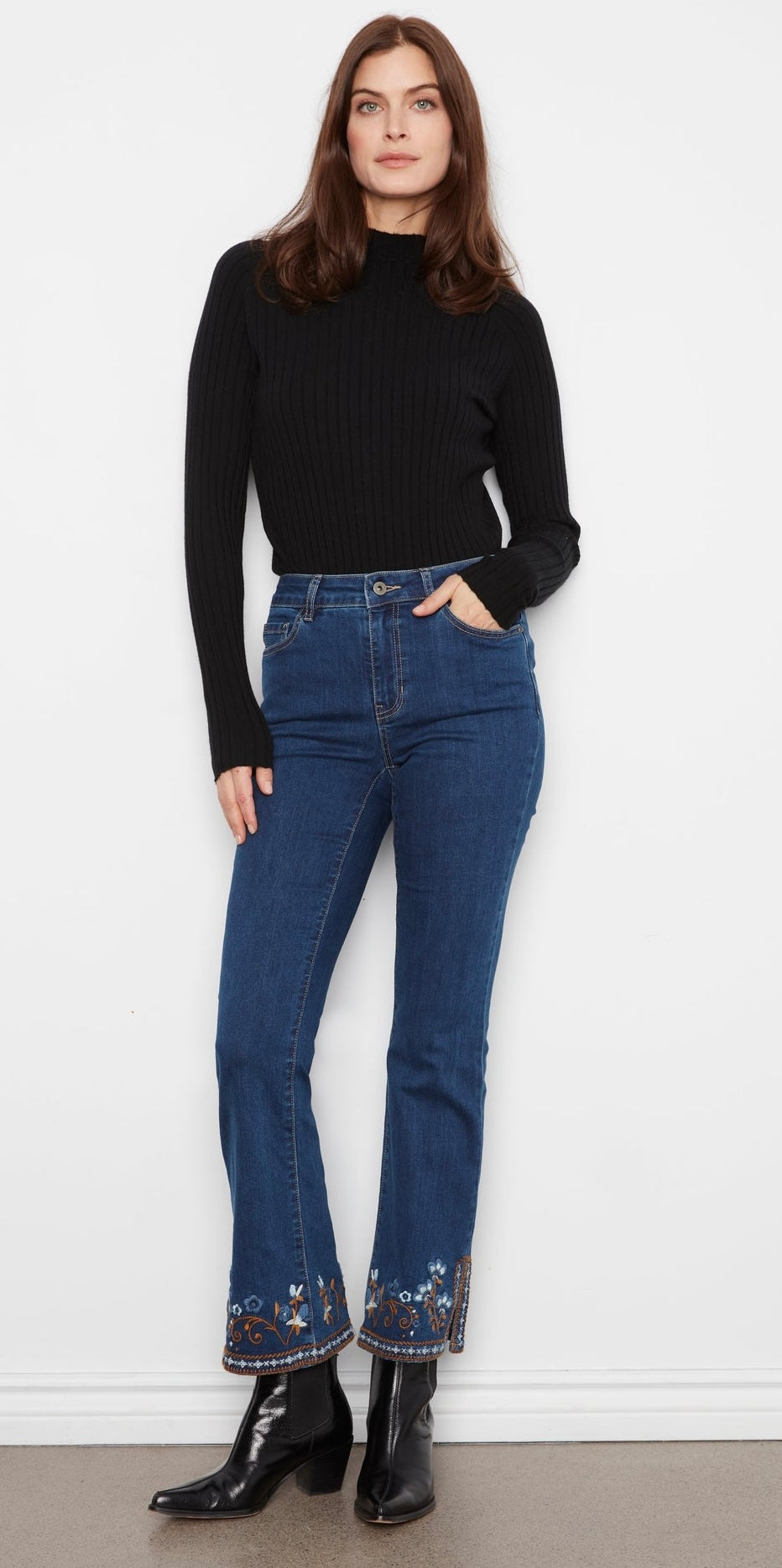 Person wearing blue jeans with floral embroidery at the bottom, standing against a white wall.
