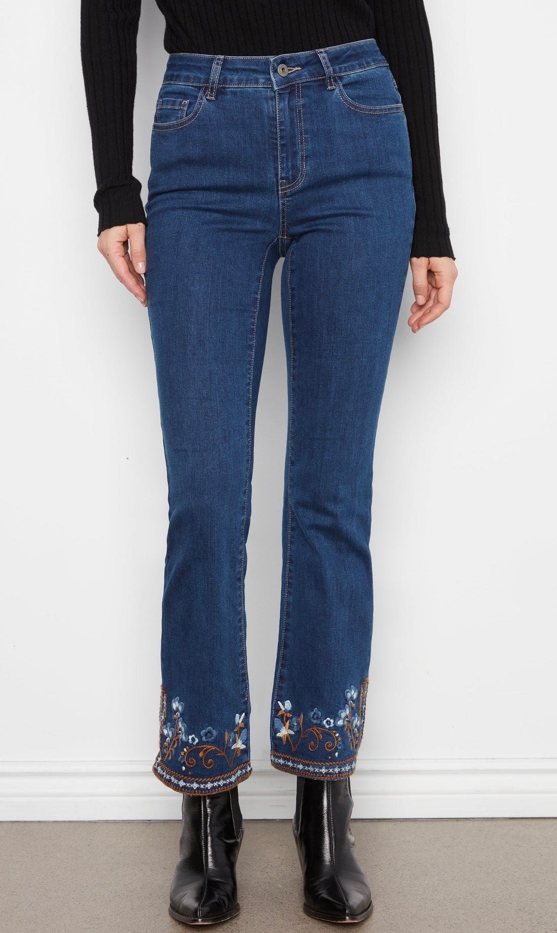 Person wearing blue jeans with floral embroidery at the bottom, standing against a white wall.