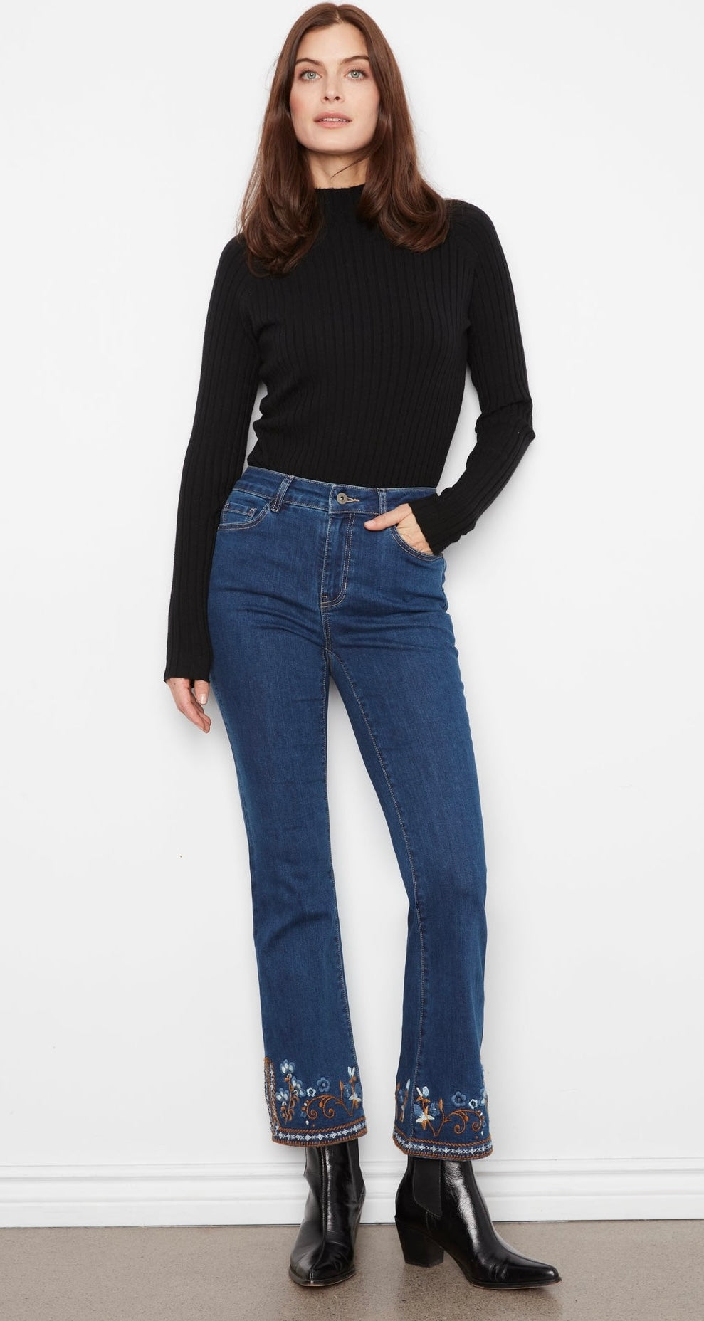 Person wearing blue jeans with floral embroidery at the bottom, standing against a white wall.