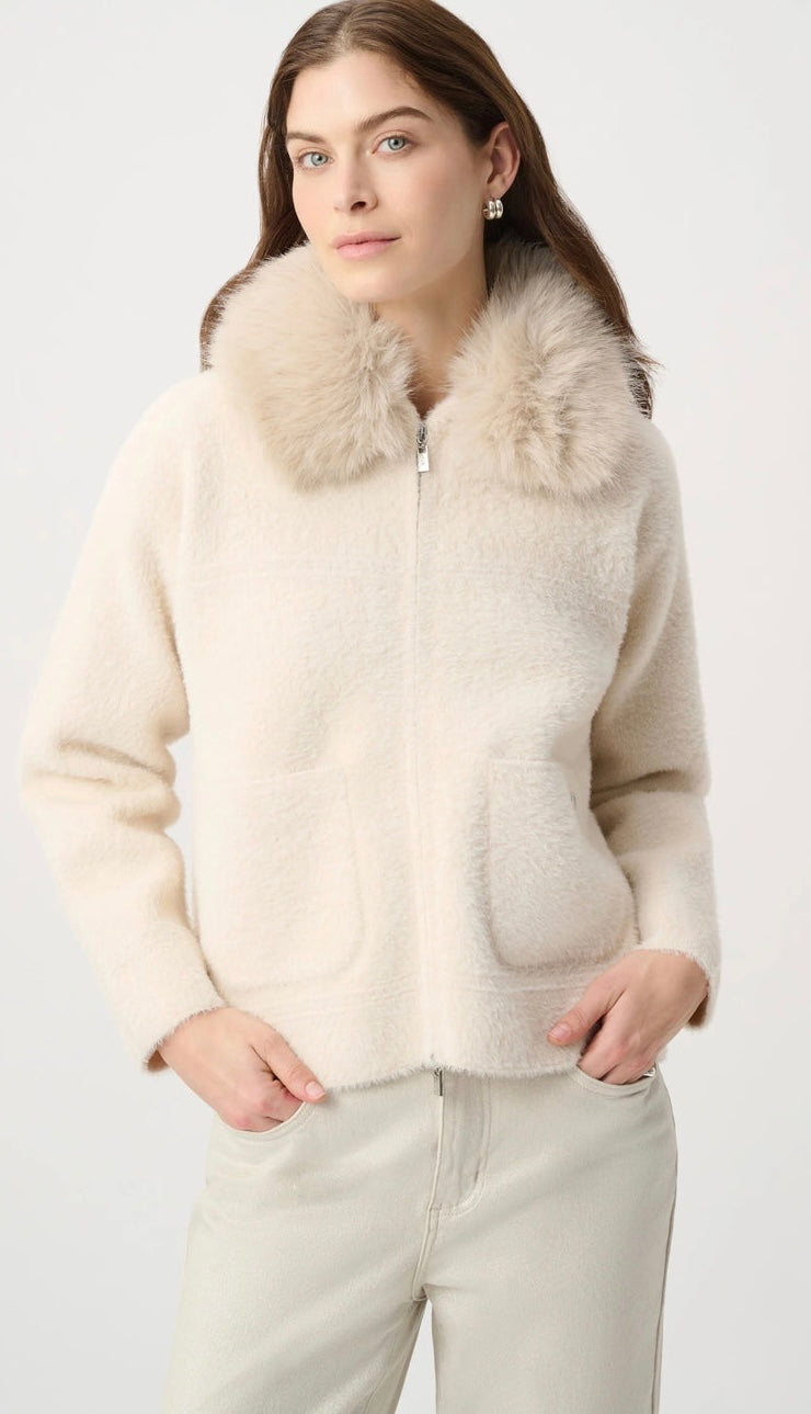 A woman wearing a cream-colored fuzzy zip-up jacket with a large faux-fur collar, paired with light beige trousers, against a plain light gray background.