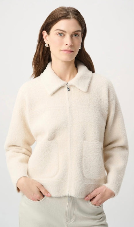 A woman wearing a cream-colored, fuzzy zip-up jacket with a collar and front pockets, paired with light beige trousers, against a plain light gray background.
