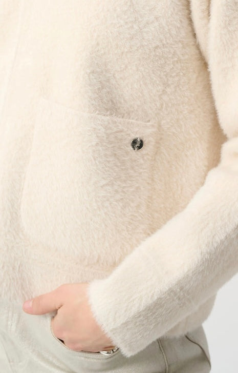 Close-up of a person wearing a soft, fuzzy cream-colored cardigan with a chest pocket and a small dark button detail, paired with light gray pants.