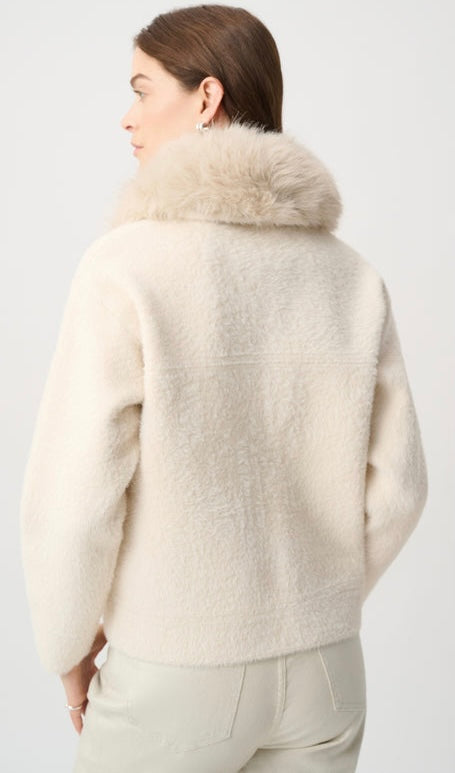 A woman shown from the back wearing a cream-colored fuzzy teddy jacket with a fluffy faux-fur collar, paired with light beige trousers, against a plain light gray studio background.
