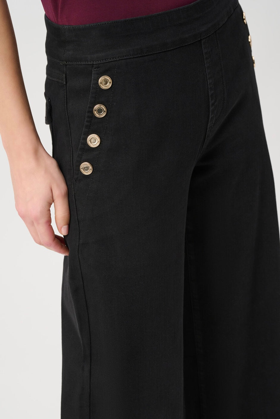 Close-up of a person wearing black wide-leg pants with a high waist and five decorative gold-tone buttons on the side, paired with a dark burgundy top against a light gray background.