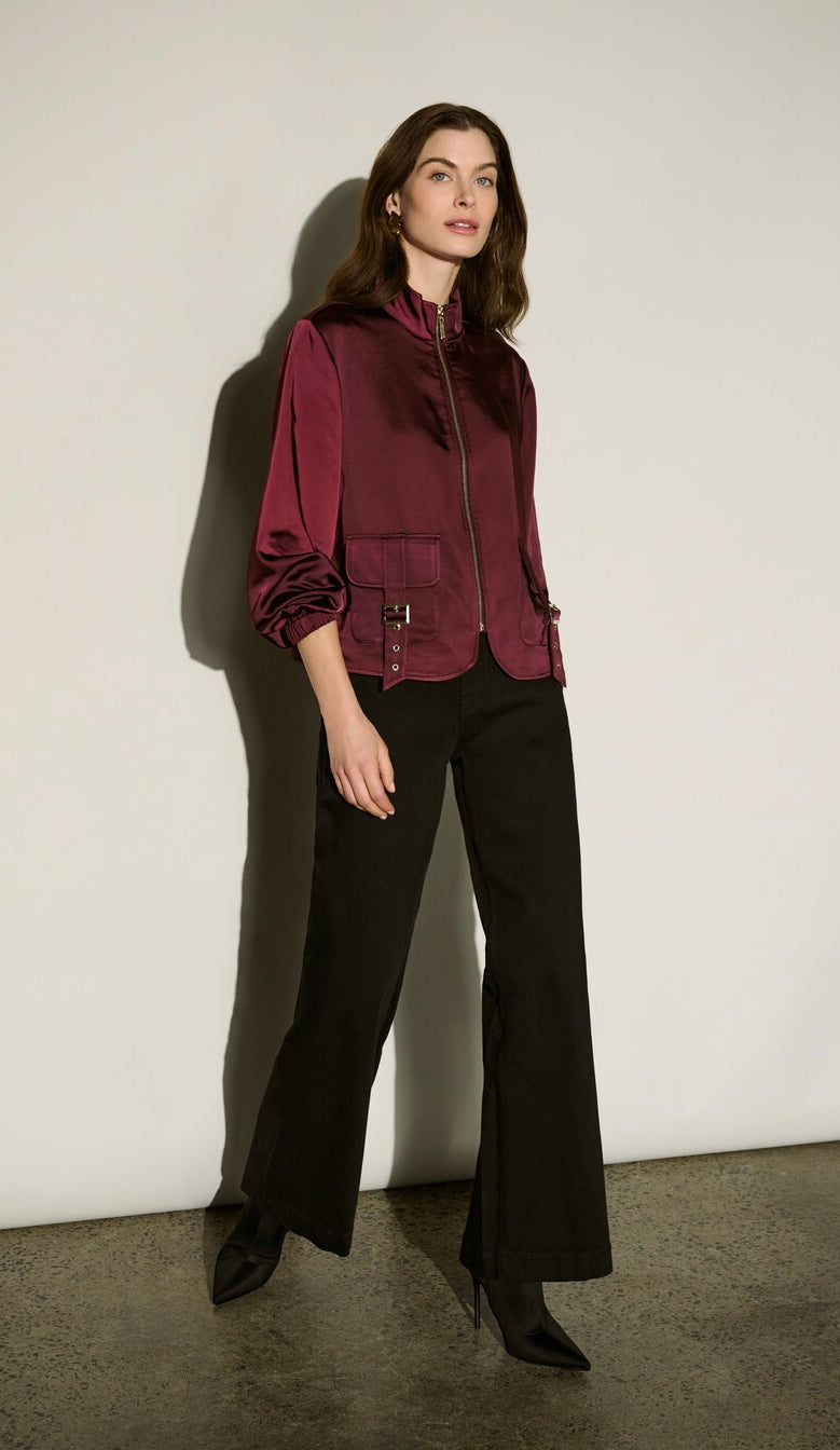 A model stands against a plain off-white wall wearing a dark burgundy satin zip-up jacket with black wide-leg trousers and black pointed-toe ankle boots.