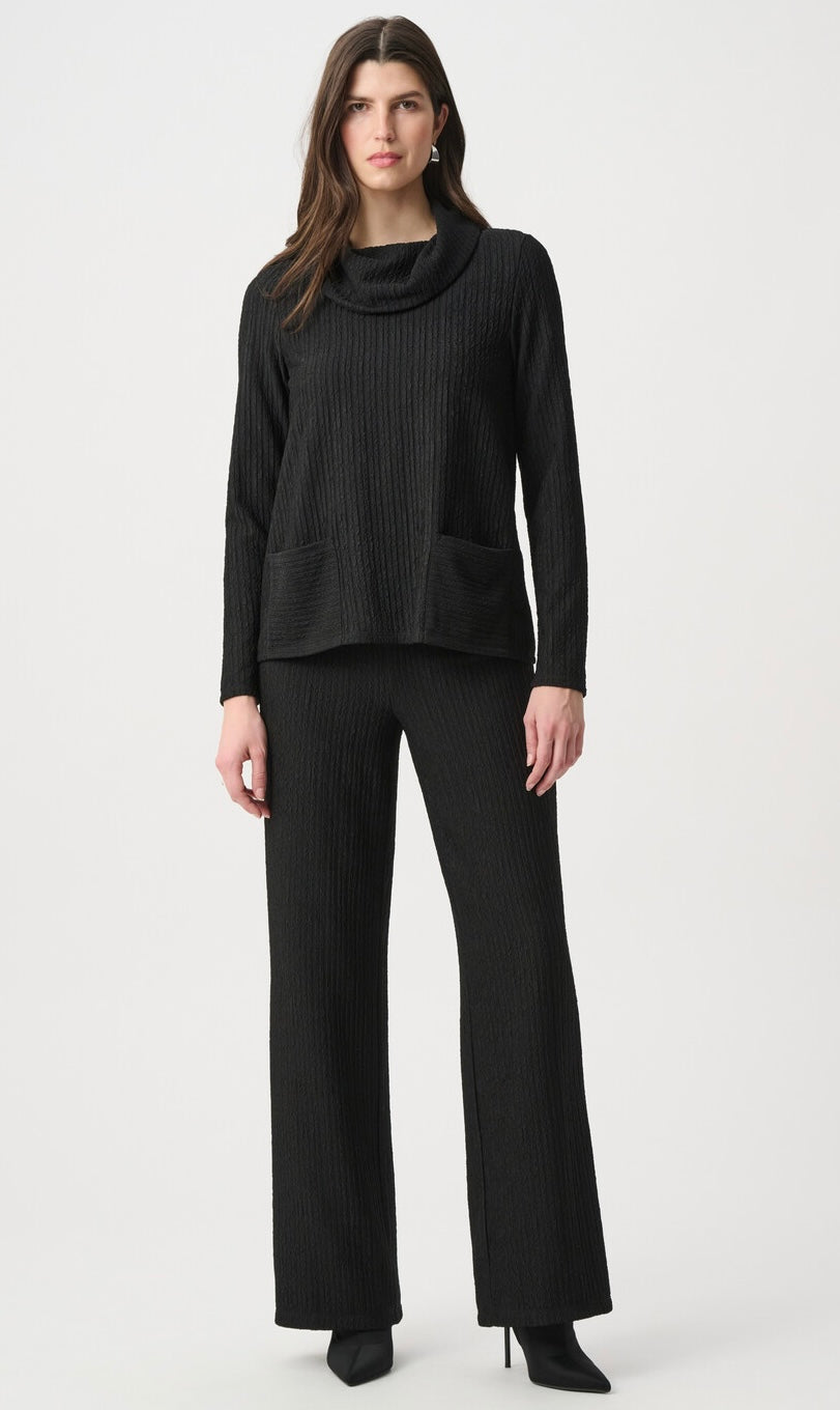 A woman modeling a black ribbed knit two-piece outfit consisting of a cowl-neck long-sleeve top and matching wide-leg trousers, photographed against a light gray studio background.