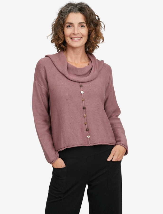 A woman wearing a dusty rose cowl-neck knit sweater with a vertical row of decorative buttons down the front, paired with black pants, against a light gray studio background.