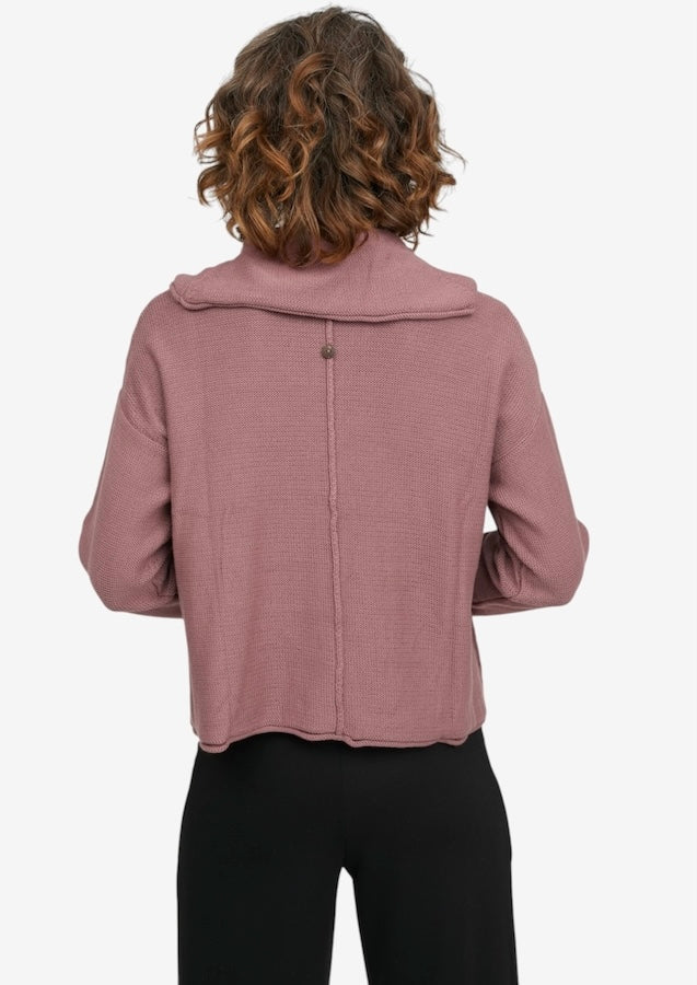 A woman viewed from the back wearing a dusty rose ribbed knit cropped jacket with a wide collar and black pants, against a plain light gray background.