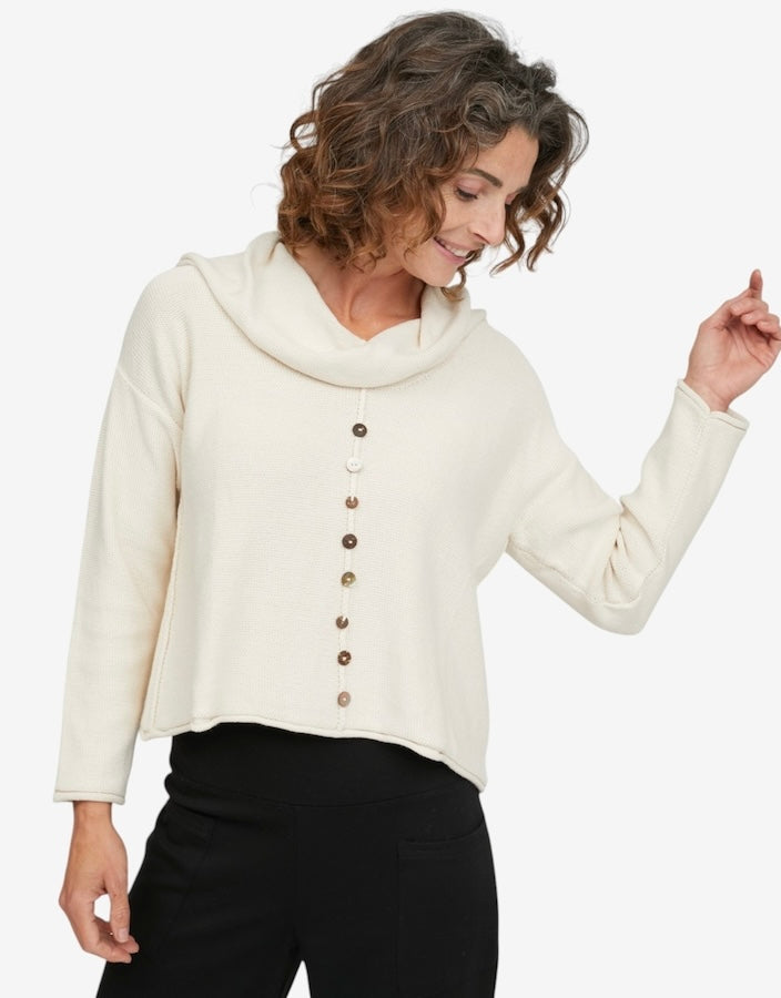 A woman wearing a cream-colored long-sleeve cowl neck top with a vertical row of brown buttons down the front, paired with black pants, against a plain white background.