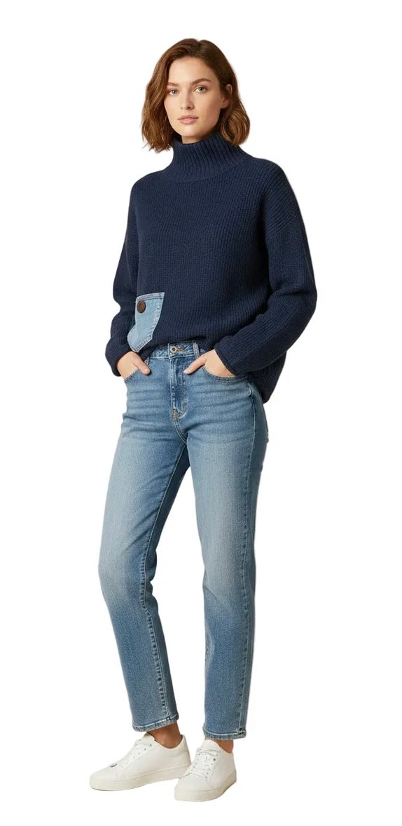 A woman modeling a navy blue turtleneck sweater with a light blue denim patch pocket on the front, paired with light-wash blue jeans and white sneakers, against a plain white background.