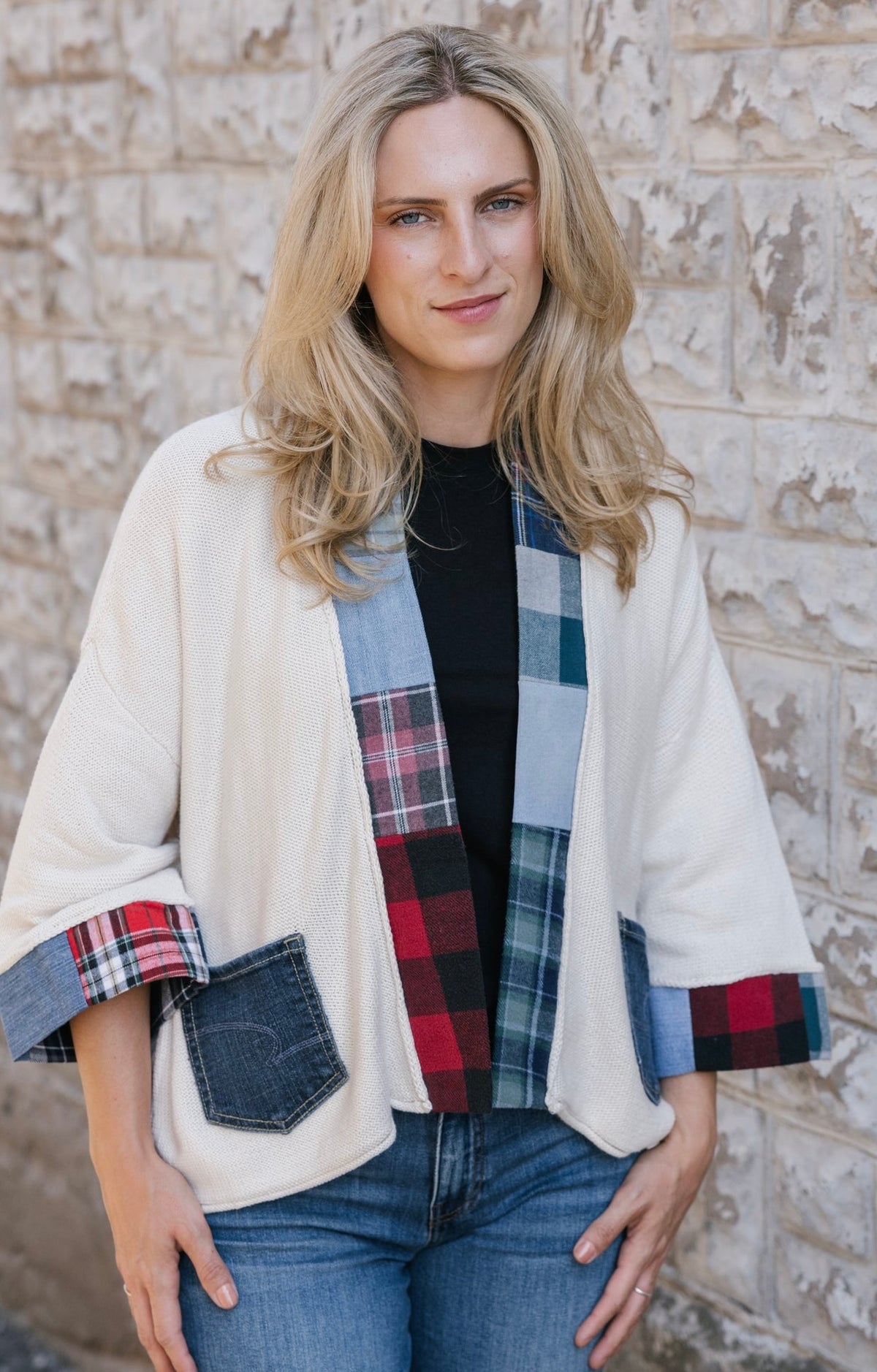 A woman wearing a cream-colored patchwork cardigan with plaid and denim details, layered over a black top and blue jeans, standing against a textured stone wall.