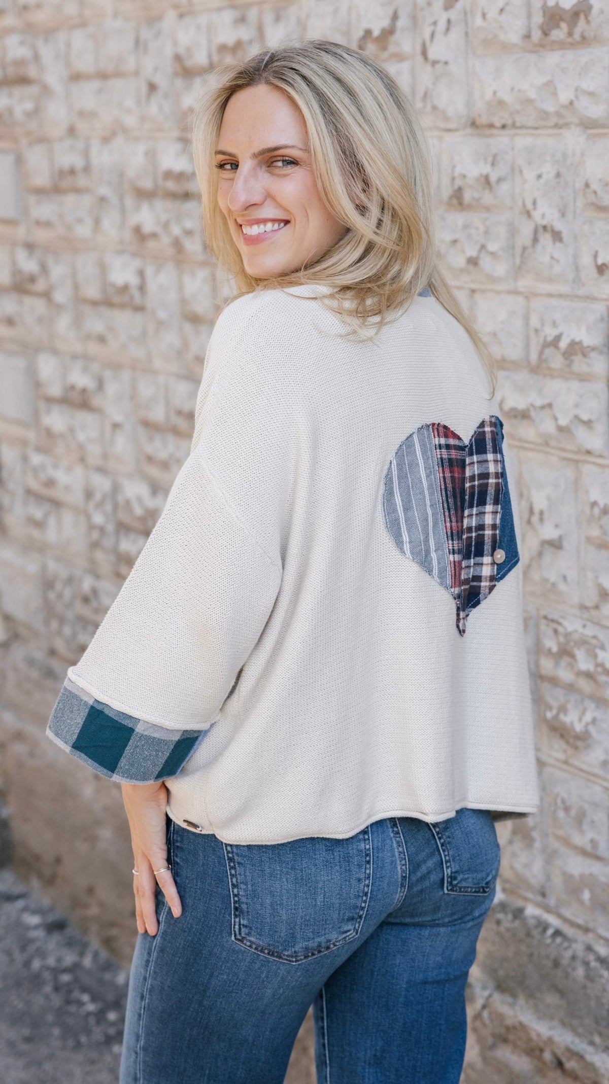 A woman wearing a cream-colored knit sweater with a blue and red plaid heart patch on the back and matching plaid trim on the sleeve cuffs, paired with blue jeans, standing against a light-colored brick wall.