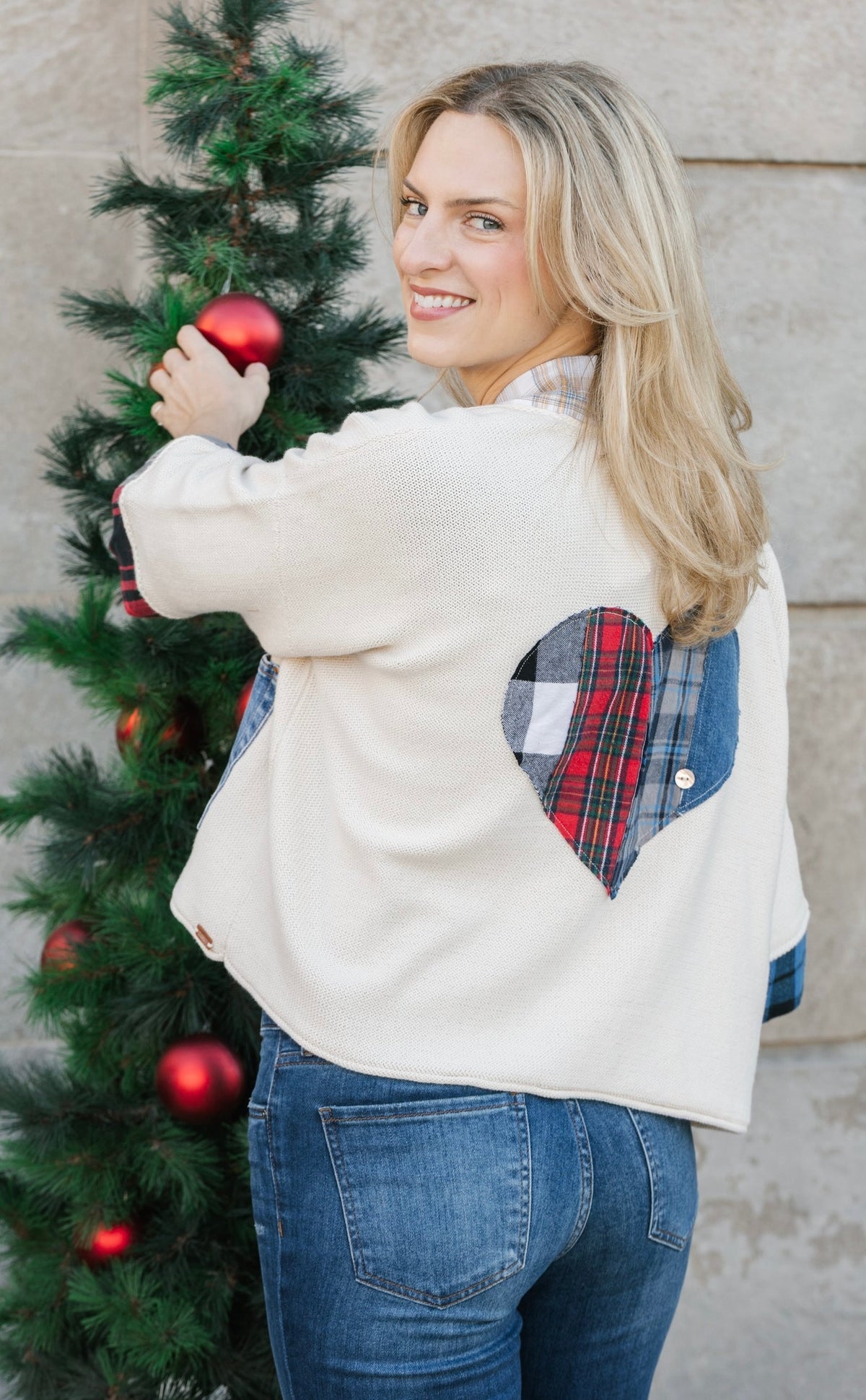 A woman wearing a cream knit sweater with a heart-shaped plaid patch on the back, standing next to a decorated Christmas tree.