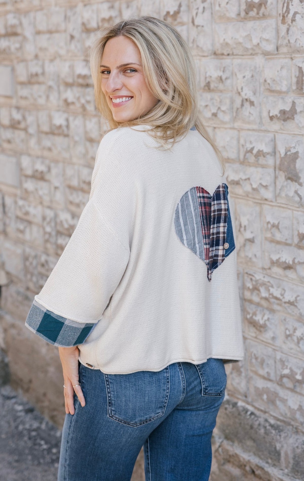 A woman wearing a cream-colored knit poncho sweater with a blue and red plaid heart patch on the back and matching plaid turned-back cuffs, standing against a light brick wall.