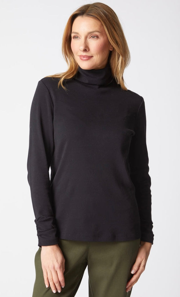 A woman wearing a black long-sleeve mock neck turtleneck top and olive green pants, standing against a light gray studio background.