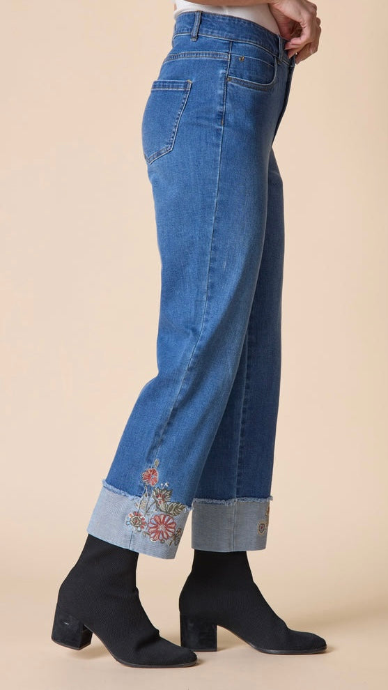 A side view of a person wearing blue cropped jeans with rolled cuffs and floral embroidery near the hem, paired with black ankle boots, against a solid beige background.
