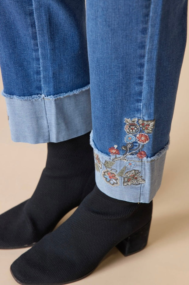 Close-up of a person's legs wearing rolled-up blue denim jeans with floral embroidery on the cuffs, paired with black knit ankle boots, against a solid beige background.