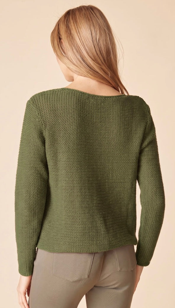 A woman shown from the back wearing a textured olive green knit sweater and light gray pants against a solid beige background.