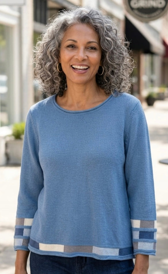 A smiling woman with curly gray hair wearing a light blue long-sleeve knit top with striped cuffs and hem, standing outdoors on a sidewalk.