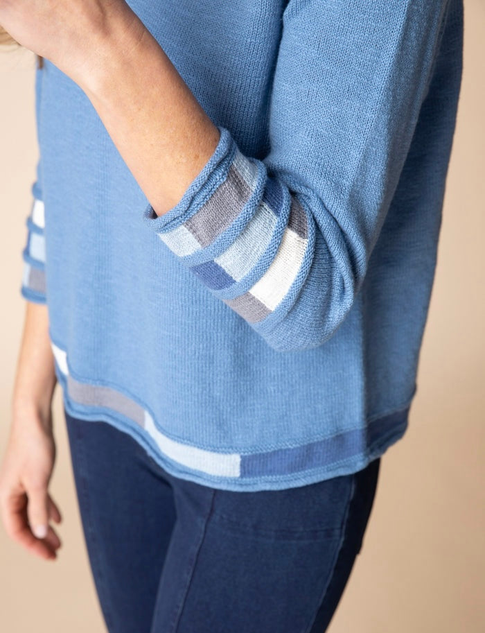 A close-up of a person wearing a light blue knit sweater with striped cuffs and hem, paired with dark blue pants, against a plain beige background.
