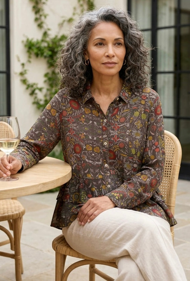 A woman with curly gray hair sits on a woven chair at an outdoor patio table, wearing a long-sleeve button-up shirt with a dense multicolor botanical print on a dark brown base and light beige trousers.