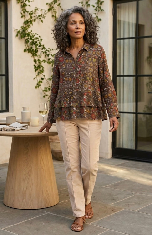 A woman stands on a patio wearing a long-sleeve button-up shirt with a dense multicolor floral and paisley print in earthy tones, paired with light beige straight-leg pants and brown sandals.