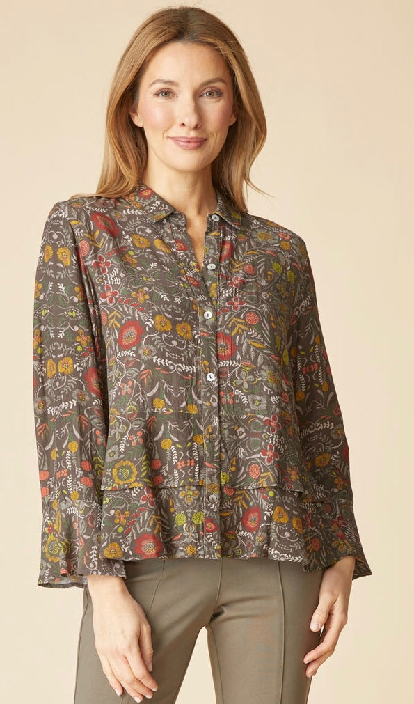 A woman modeling a long-sleeve button-up blouse with an intricate multicolor floral and paisley print on a brown base, paired with olive green pants, set against a solid beige background.