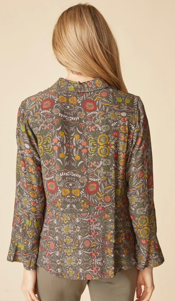 A woman shown from the back wearing a long-sleeve collared blouse with an all-over floral and botanical print in muted olive, red, yellow, and orange tones, paired with light olive pants against a plain beige studio background.