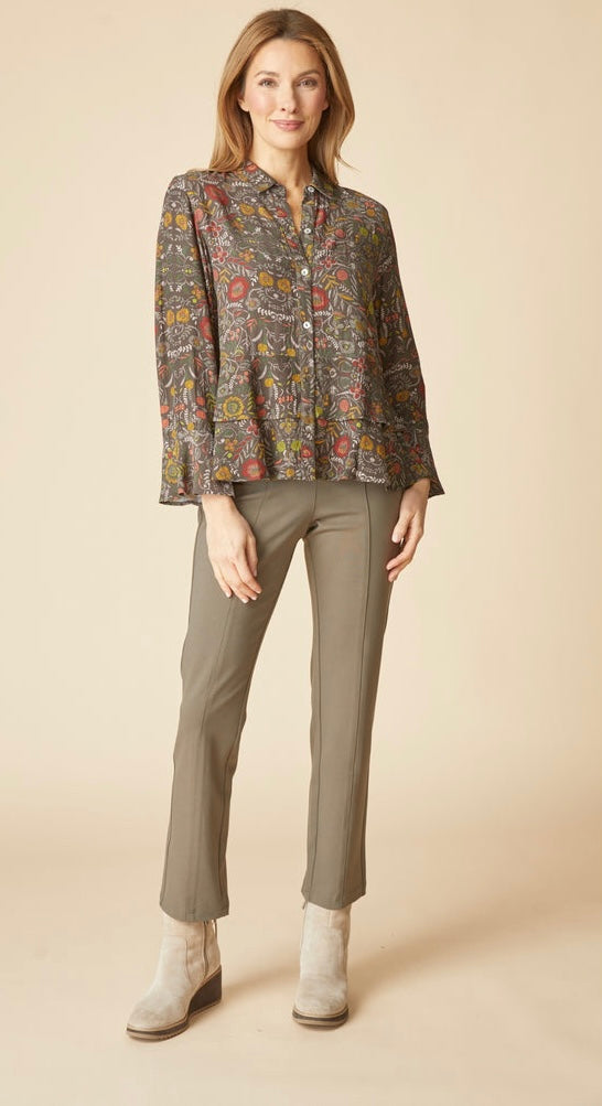 A woman modeling a long-sleeve button-up blouse with a multicolor abstract print on a brown base, paired with olive green pants and light beige ankle boots, against a solid beige studio background.