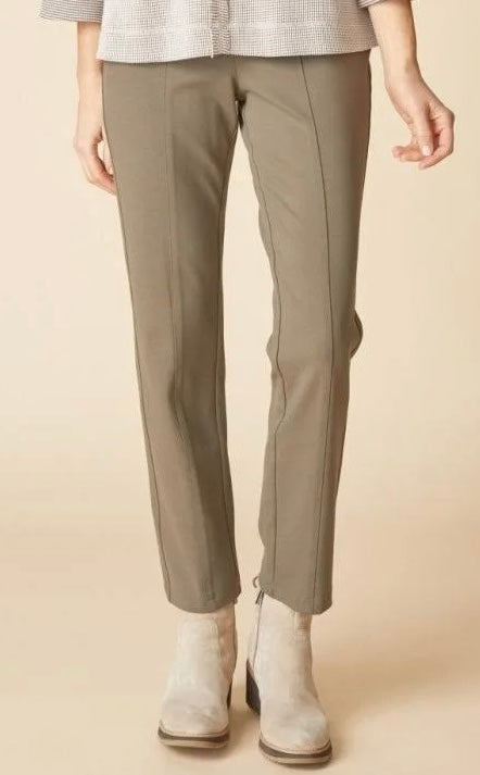 A person wearing slim-fit cropped trousers in a muted taupe-gray color, paired with a light patterned top and light-colored ankle boots, against a solid beige studio background.