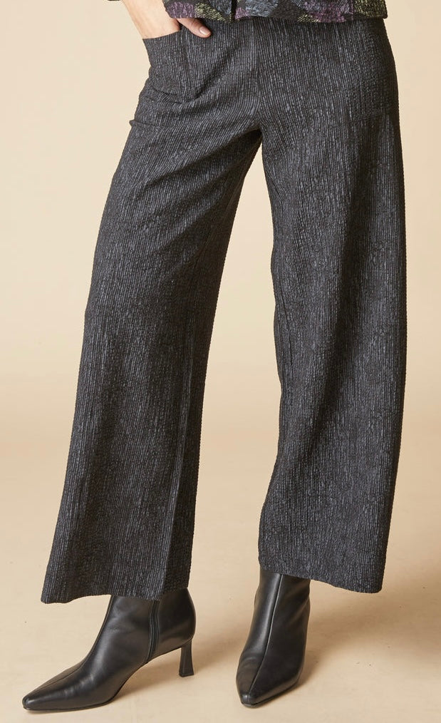 A person wearing dark charcoal gray wide-leg textured pants and black pointed-toe ankle boots, standing against a plain beige studio background.