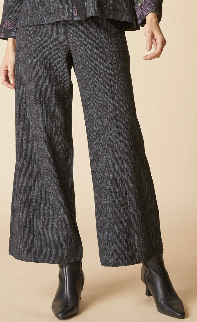 A person wearing dark charcoal wide-leg cropped trousers with a subtle heathered texture, styled with black pointed-toe ankle boots against a solid beige studio background.