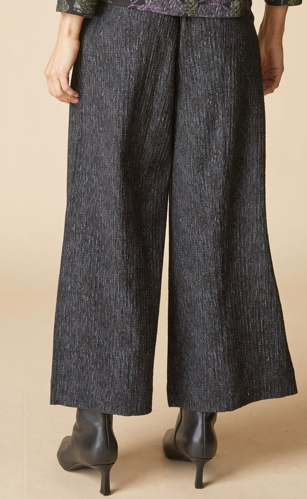 A person shown from the back wearing dark charcoal wide-leg cropped pants and black ankle boots against a plain beige studio background.