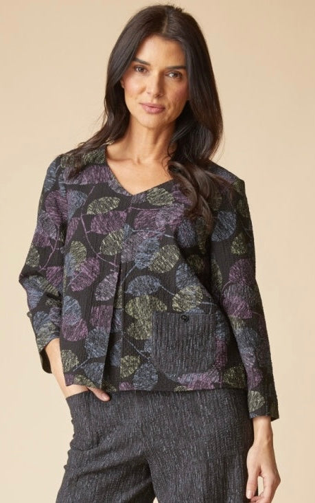 A woman modeling a dark patterned long-sleeve top with purple, green, and gray leaf motifs, paired with dark textured pants, against a solid beige background.