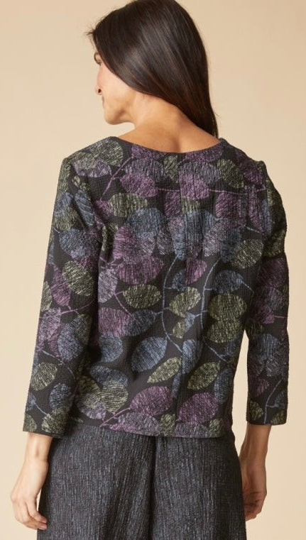 A woman shown from the back wearing a dark blouse with a purple and olive green leaf pattern, paired with dark gray textured pants, against a solid beige background.