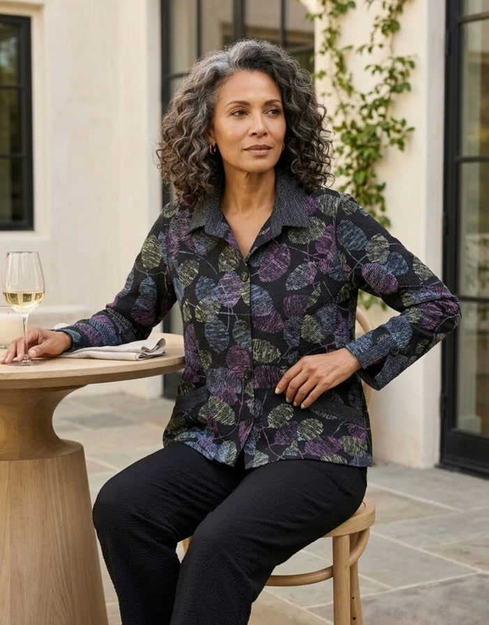 A woman with curly gray hair sits on a wooden chair at an outdoor patio table, wearing a long-sleeve button-up shirt with a dark navy and purple botanical leaf pattern, paired with black pants.