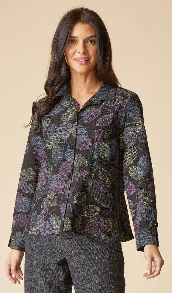A woman modeling a long-sleeve button-up shirt with a dark charcoal base and an abstract leaf-like pattern in purple, olive green, and gray, paired with textured gray pants against a solid beige background.