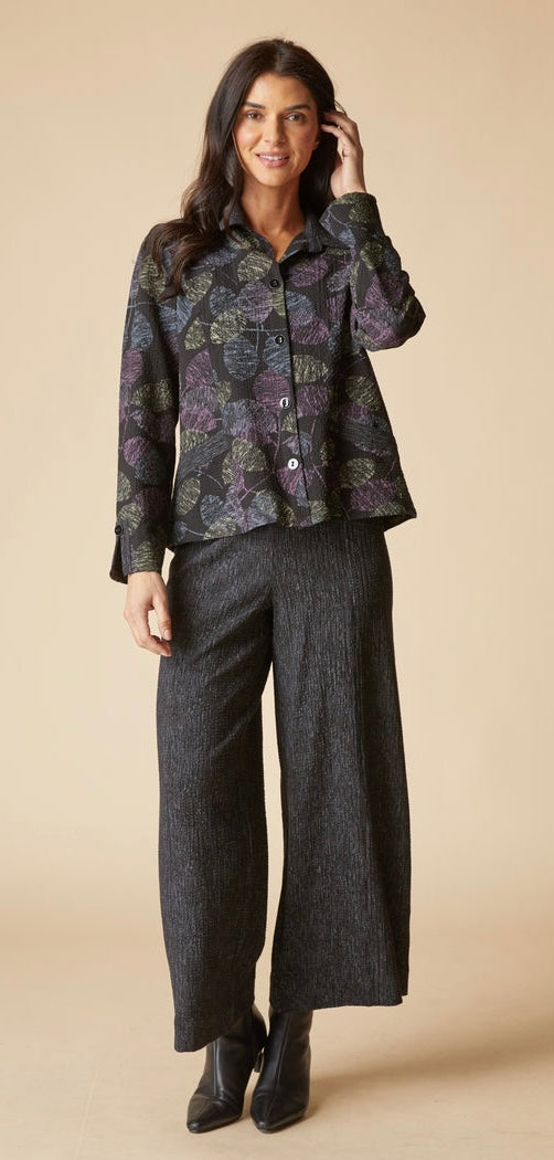 A woman modeling a long-sleeve button-up shirt with a dark purple and green floral pattern, paired with dark charcoal wide-leg trousers and black ankle boots, against a solid beige studio background.