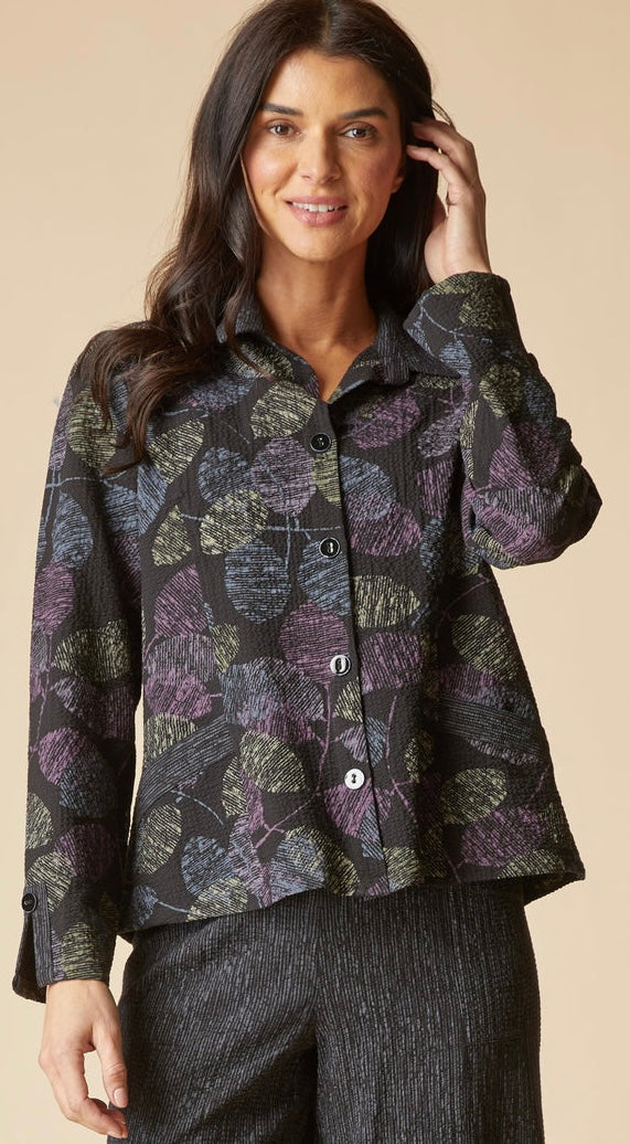 A woman wearing a long-sleeve button-front collared shirt with an abstract leaf-like pattern in black, purple, and olive green, paired with dark textured pants, against a solid beige background.
