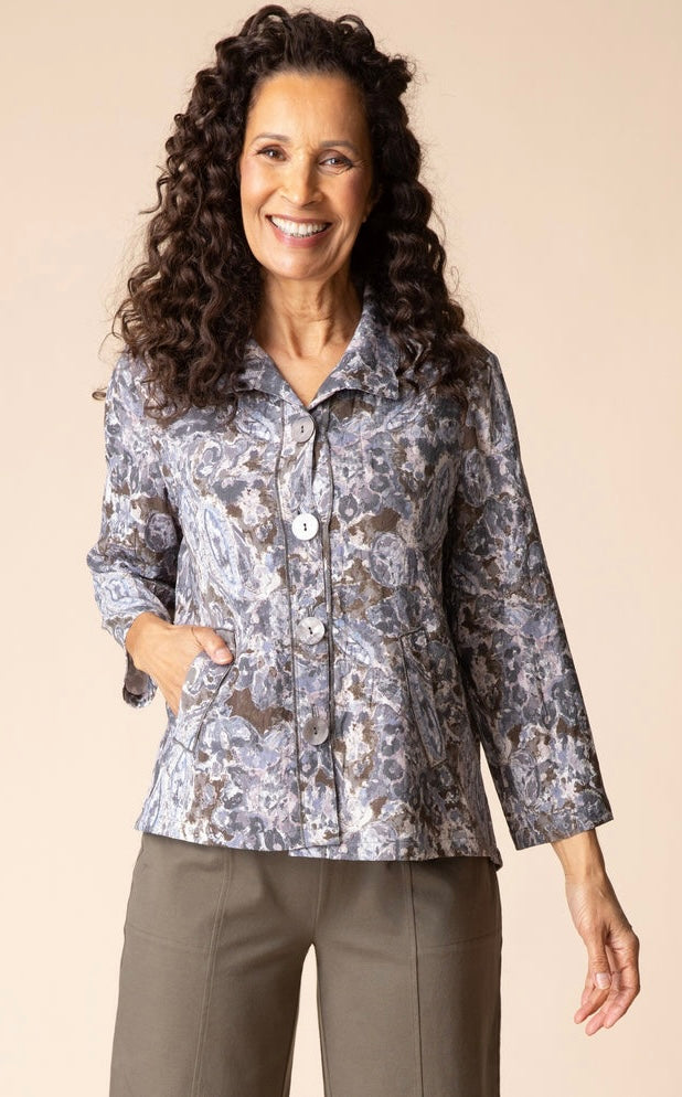 A smiling woman with long curly dark hair models a long-sleeve button-front blouse with a gray, white, and brown paisley pattern, paired with taupe pants, against a solid beige studio background.