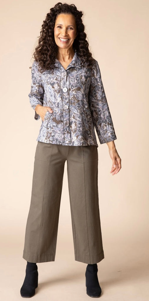 A smiling woman with long curly brown hair wearing a patterned button-up blouse with three-quarter sleeves and brown pants, standing against a solid beige studio background.