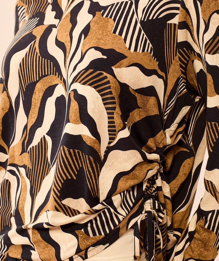 A long-sleeve knit top with a bold abstract botanical pattern in black, tan, and cream, held up on a hanger by a person in a clothing store.