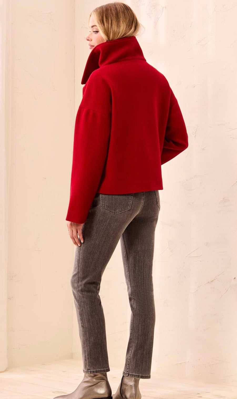 A woman shown from the back wearing a bright red cropped turtleneck jacket, gray skinny jeans, and metallic silver ankle boots, standing against a light beige textured wall. - OLIVIA'S