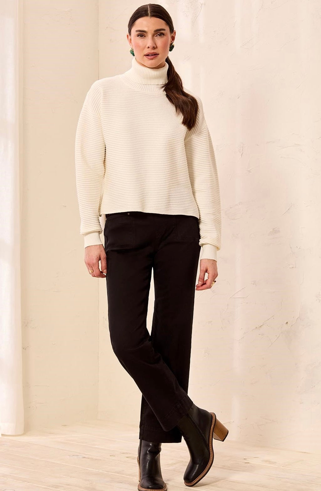 A woman modeling a cream ribbed turtleneck sweater and black pants in a warm, neutral indoor setting. - OLIVIA'S