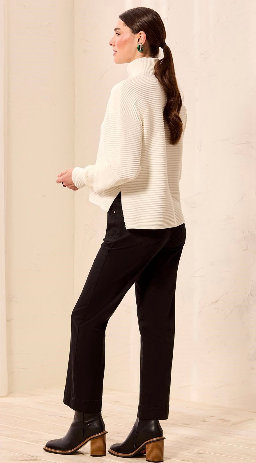 A woman wearing a cream ribbed turtleneck sweater, black trousers, and black ankle boots with wooden block heels, shown from a side-back angle against a neutral cream-colored wall. - OLIVIA'S